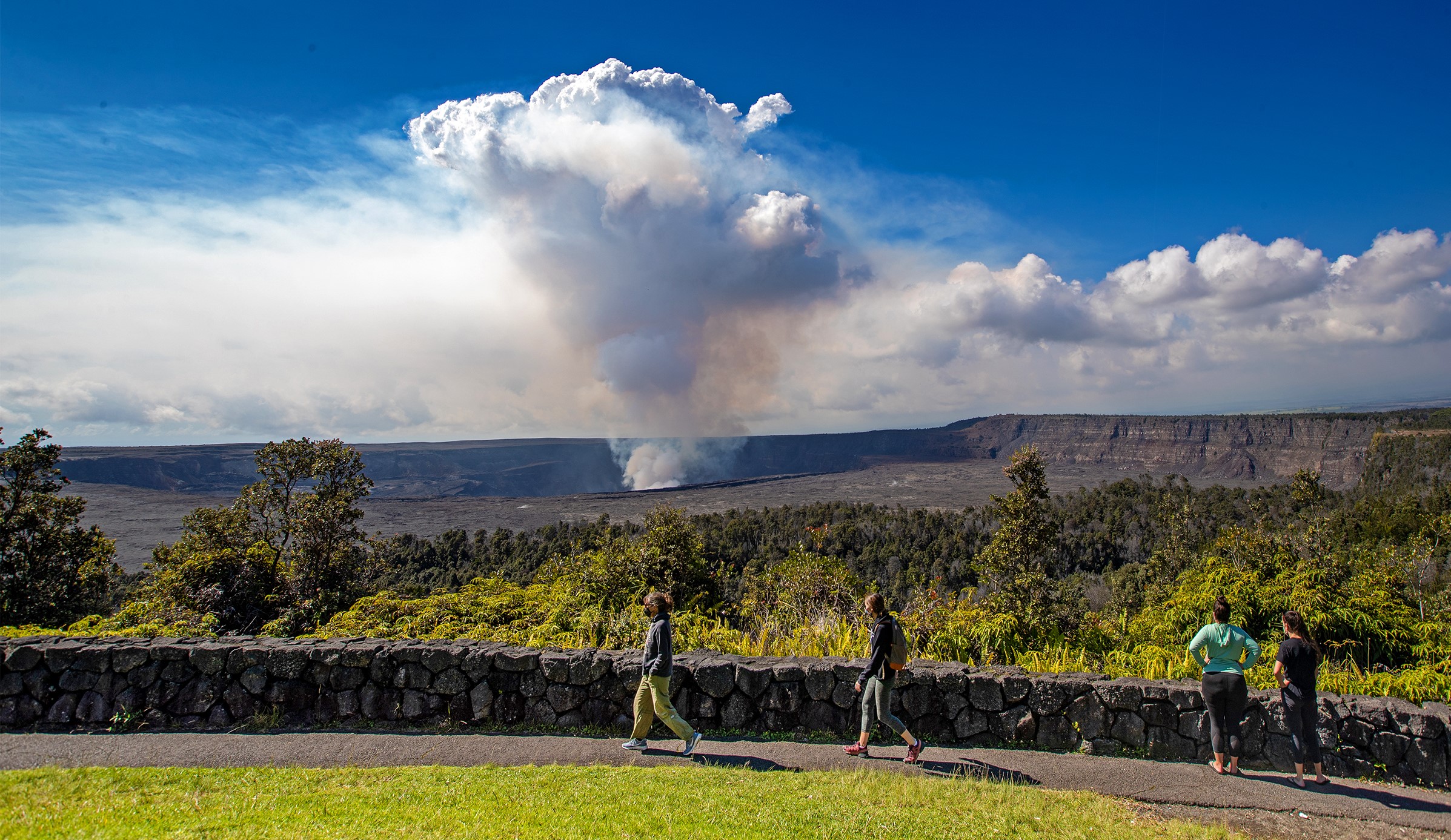 A real photograph of a roadside overlook along Crater Rim Drive in Hawaiʻi Volcanoes National Park, with a stone wall viewpoint and a sweeping volcanic landscape beyond