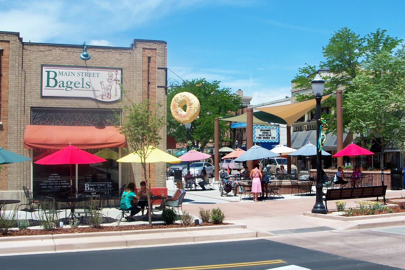 A real photograph of a shaded cafe patio in downtown Grand Junction, Colorado with people sitting at small tables and bicycles parked nearby