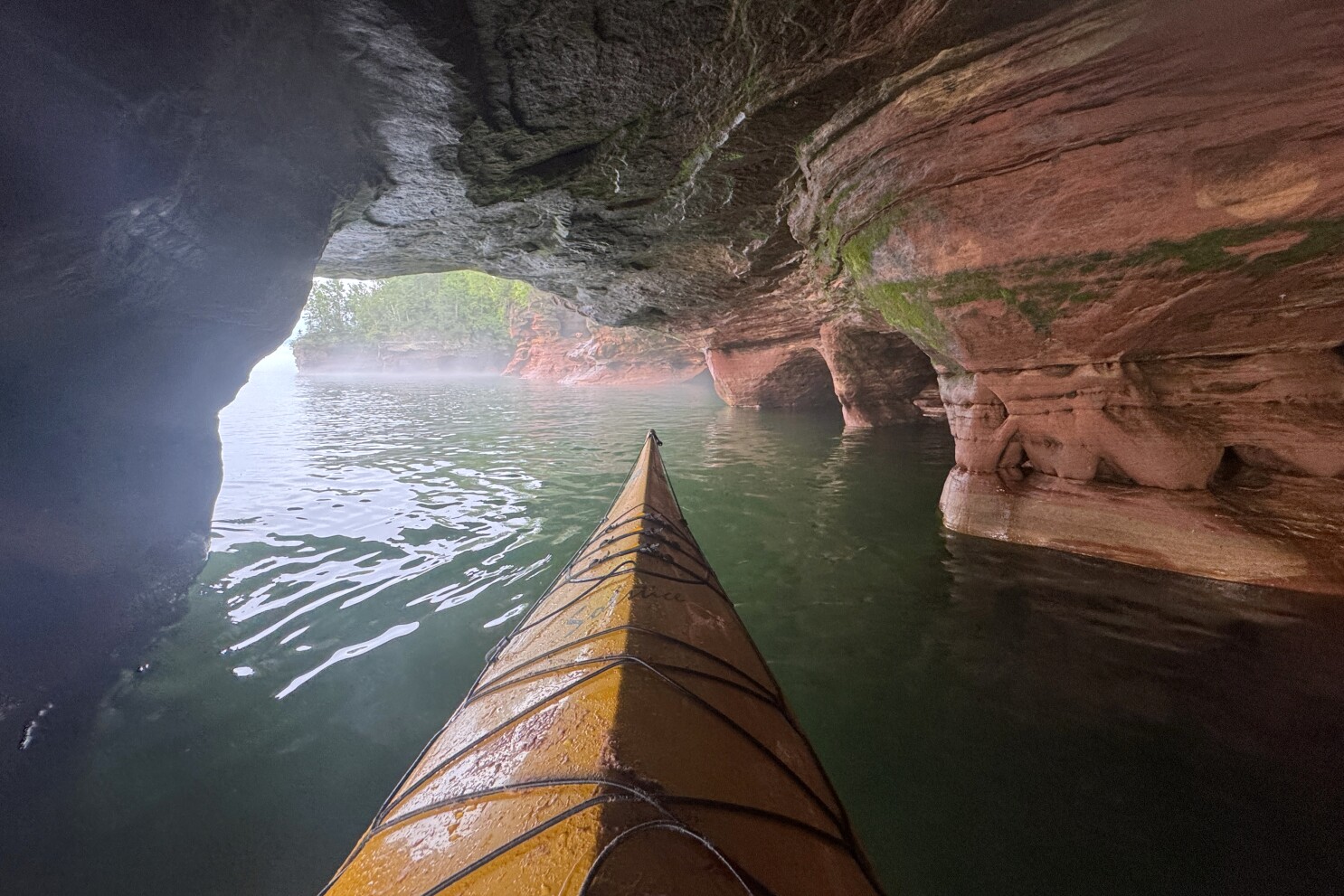 A real photograph of a single kayaker in a bright kayak approaching the entrance of a sandstone sea cave on Lake Superior, with textured rock walls and gentle waves at the cave mouth
