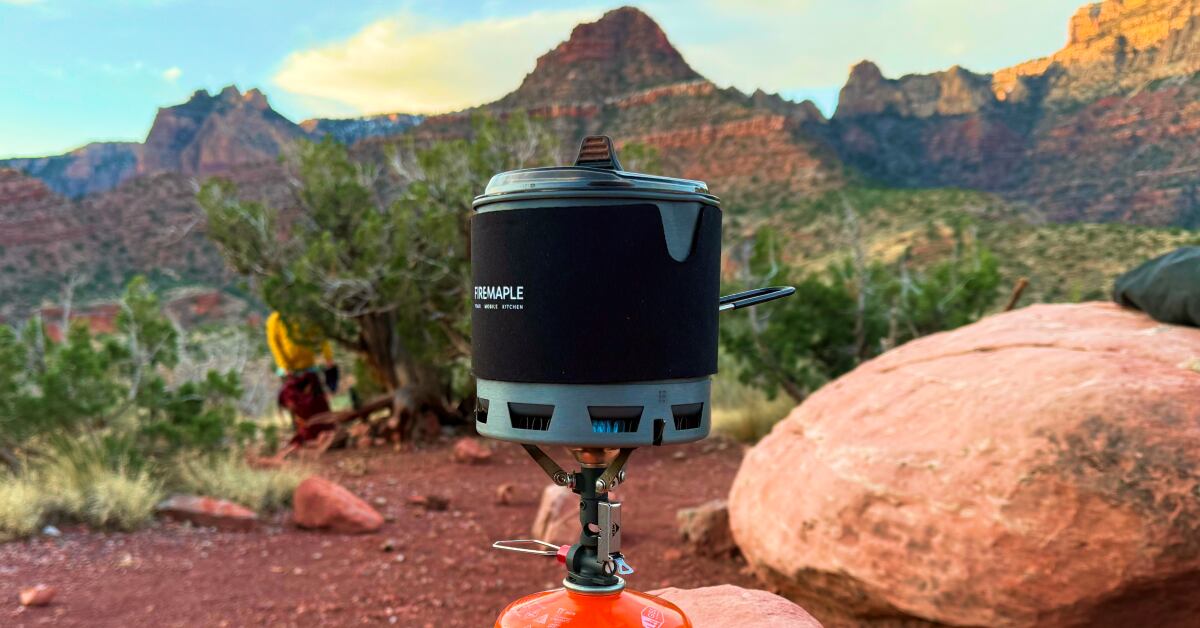 A real photograph of a small backpacking stove boiling water in a titanium pot on a flat rock near a campsite, with a canister fuel stove attached, dusk light