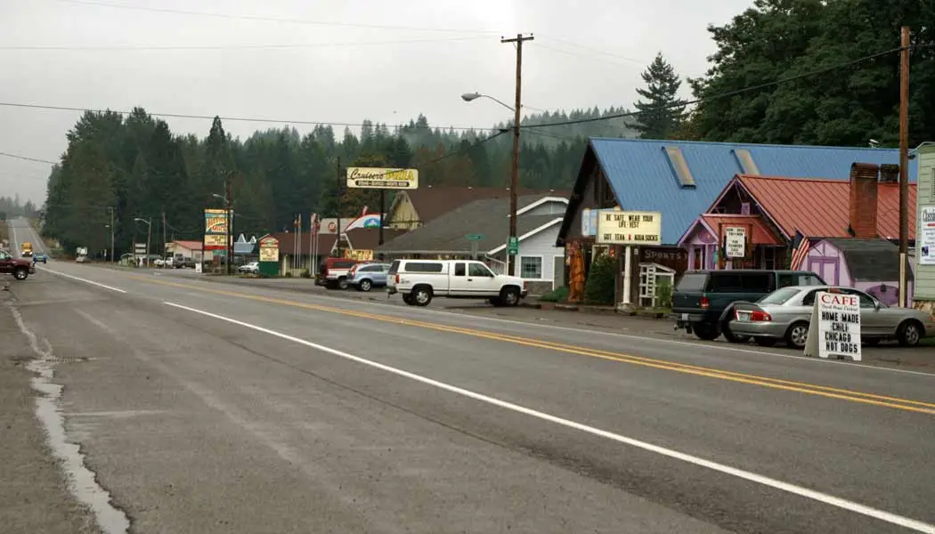 A real photograph of a small-town street in Packwood, Washington in the evening with storefronts, parked cars, and warm light reflecting off wet pavement
