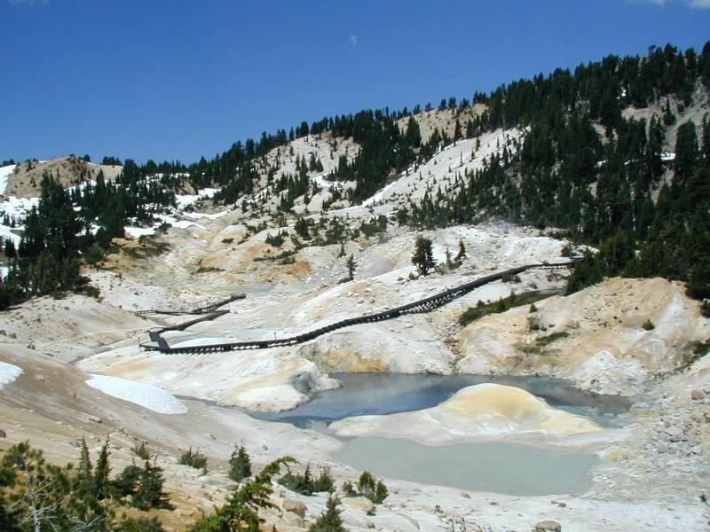 A real photograph of a steaming hydrothermal vent in Lassen Volcanic National Park with mineral stained ground and a clear blue sky, taken from a safe viewpoint