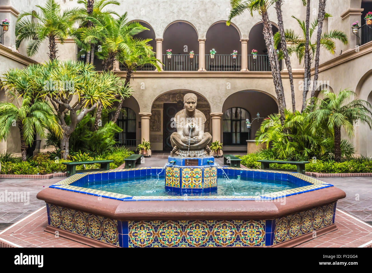 A real photograph of a sunlit courtyard in Balboa Park in San Diego, showing Spanish Colonial Revival buildings, arches, and palm trees