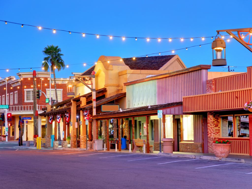 A real photograph of a sunny Old Town Scottsdale street with low adobe-style buildings, pedestrians in casual clothes, and desert plants in planters