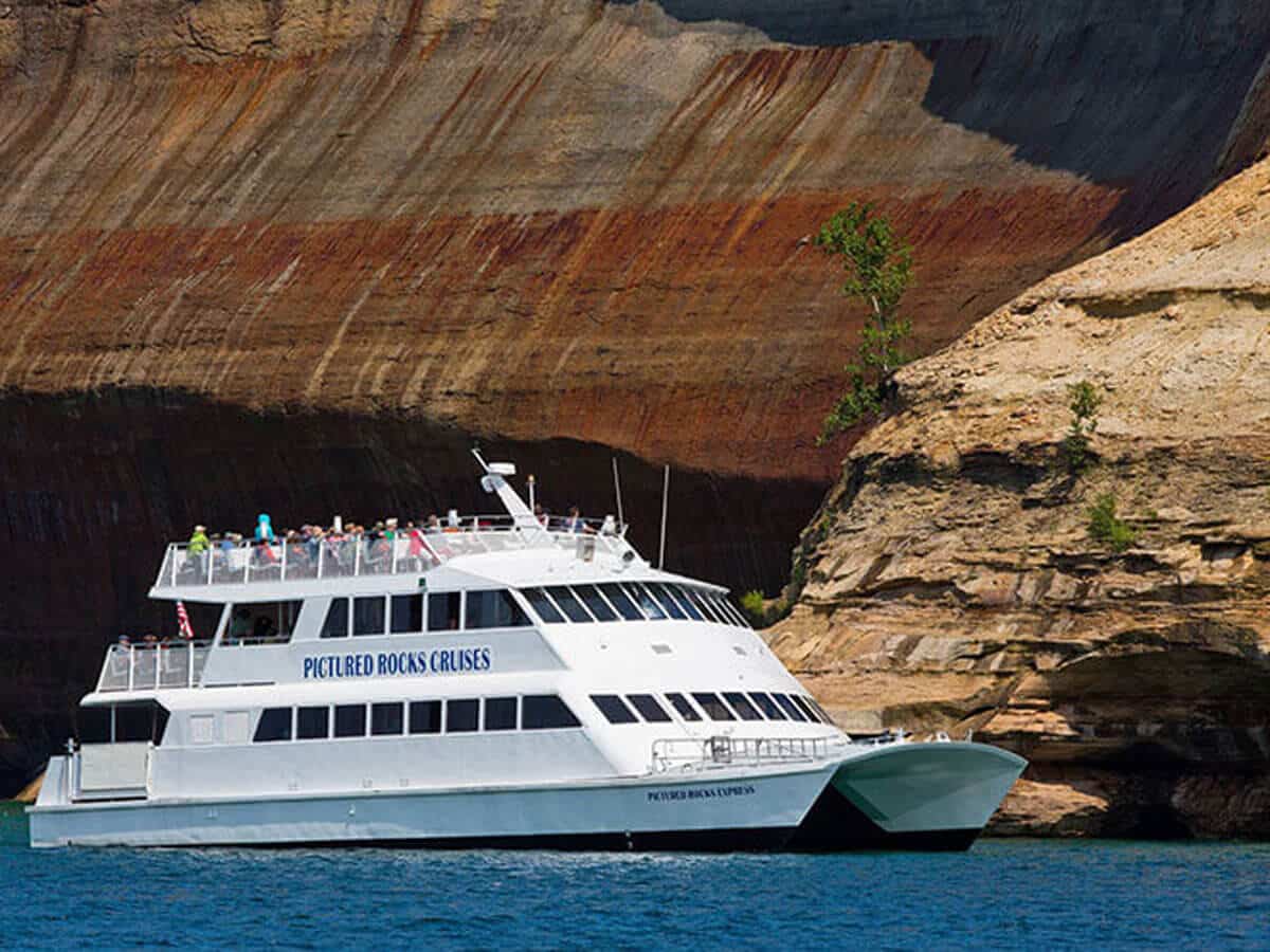 A real photograph of a tour boat departing Munising, Michigan on a sunny summer day, with passengers on the open deck and the Lake Superior shoreline in the distance