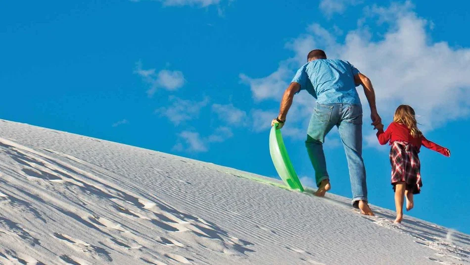 A real photograph of a traveler sitting on a waxed plastic sand sled at the top of a white gypsum dune in White Sands National Park, ready to slide down