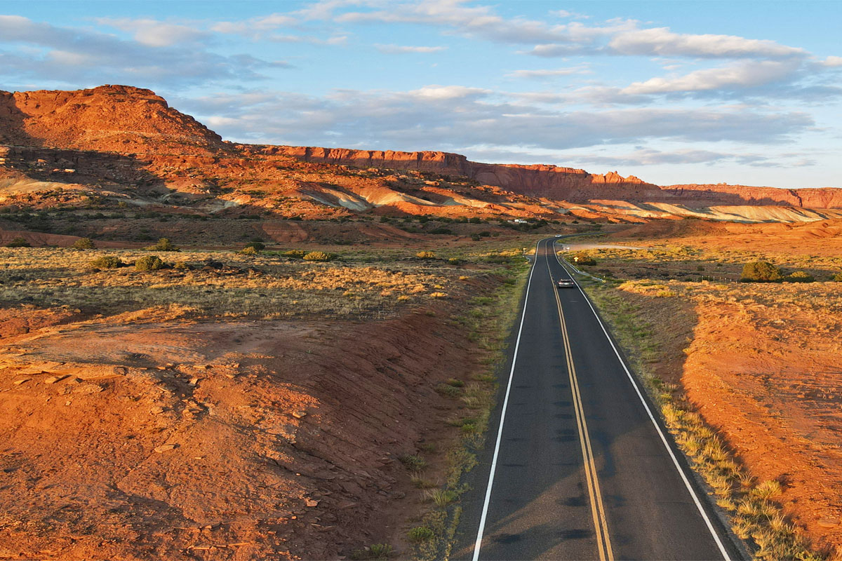 A real photograph of a two-lane Utah desert highway stretching toward distant red rock mesas, with sparse scrub on both sides and wide open sky in late afternoon light