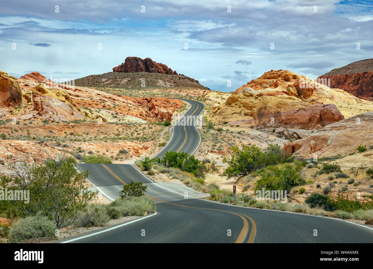 A real photograph of a two-lane road winding through Valley of Fire State Park in Nevada, with red rock hills on both sides and a clear sky above