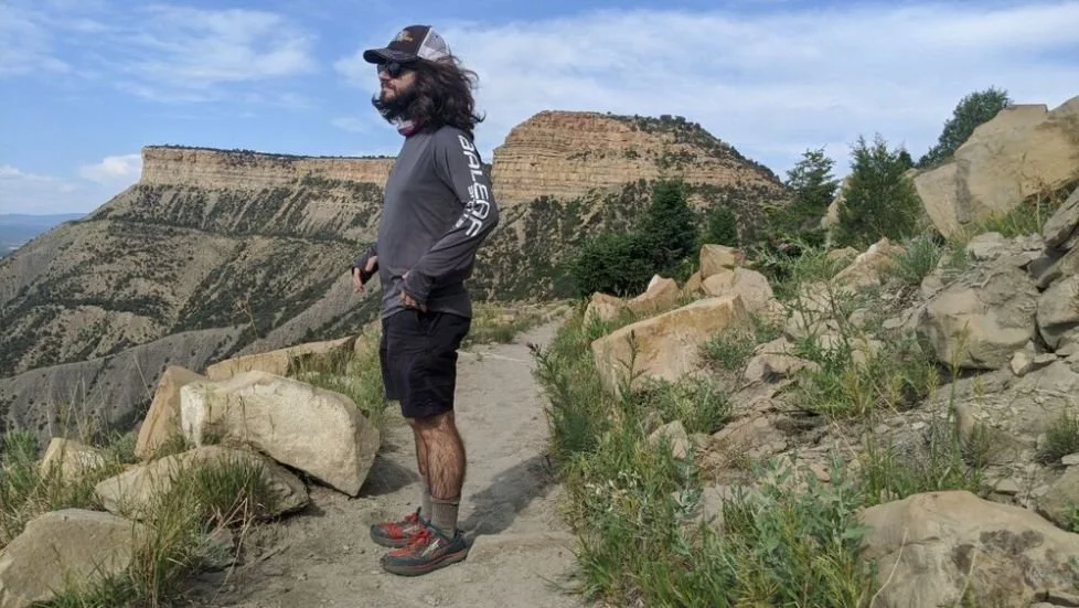 A real photograph of a visitor wearing a wide-brim sun hat and small daypack walking on a dusty trail at Mesa Verde National Park with canyon views in the background