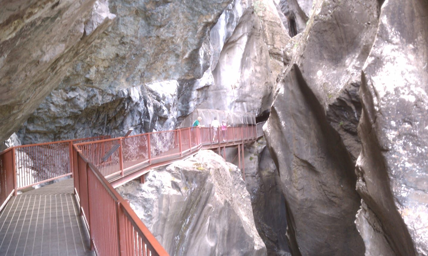 A real photograph of a waterfall crashing through the Ouray Box Canyon with a metal walkway and steep rock walls, mist in the air