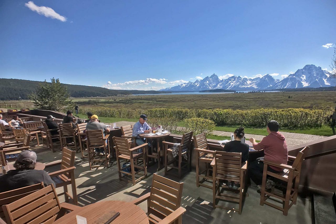 A real photograph of an inviting restaurant patio in Jackson, Wyoming at golden hour with diners seated at wooden tables and mountains faintly visible in the background