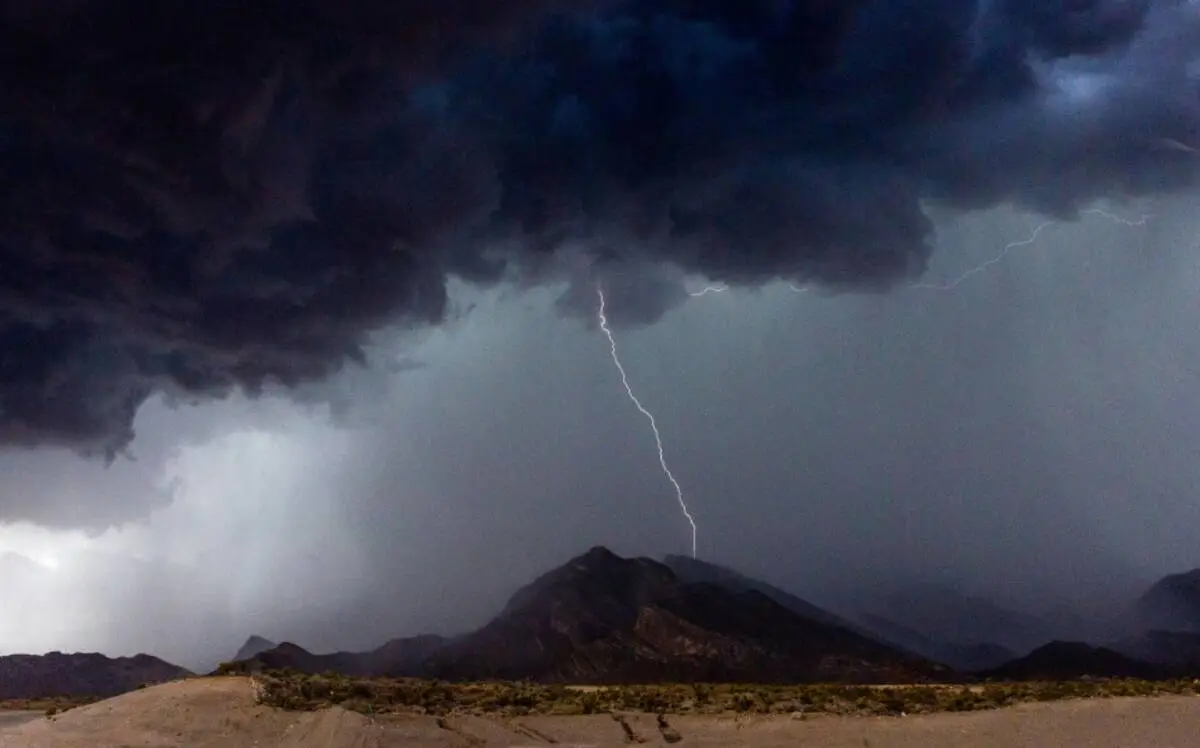 A real photograph of dark summer thunderstorm clouds building over the Mount Charleston area with sunlight still hitting the pine-covered slopes in the foreground