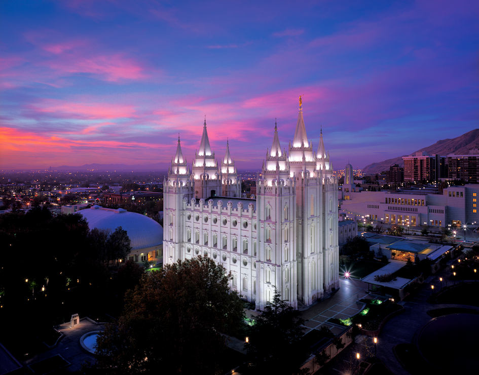 A real photograph of downtown Salt Lake City near Temple Square on a clear day, with wide sidewalks, historic stone buildings, and the Wasatch Mountains visible in the distance