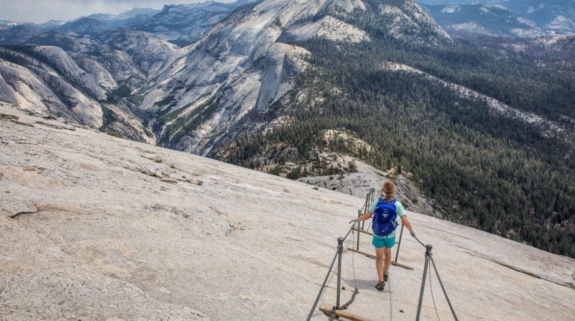 A real photograph of hikers ascending the Half Dome cables on a clear summer morning in Yosemite National Park, with gloves on and the Valley visible far below