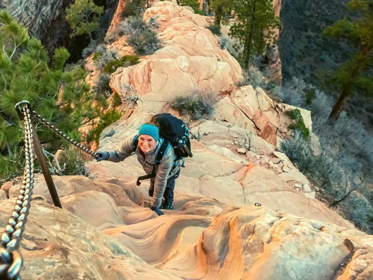A real photograph of hikers carefully holding the chains on the narrow Angels Landing ridge in Zion National Park, steep drop-offs on both sides under clear daylight