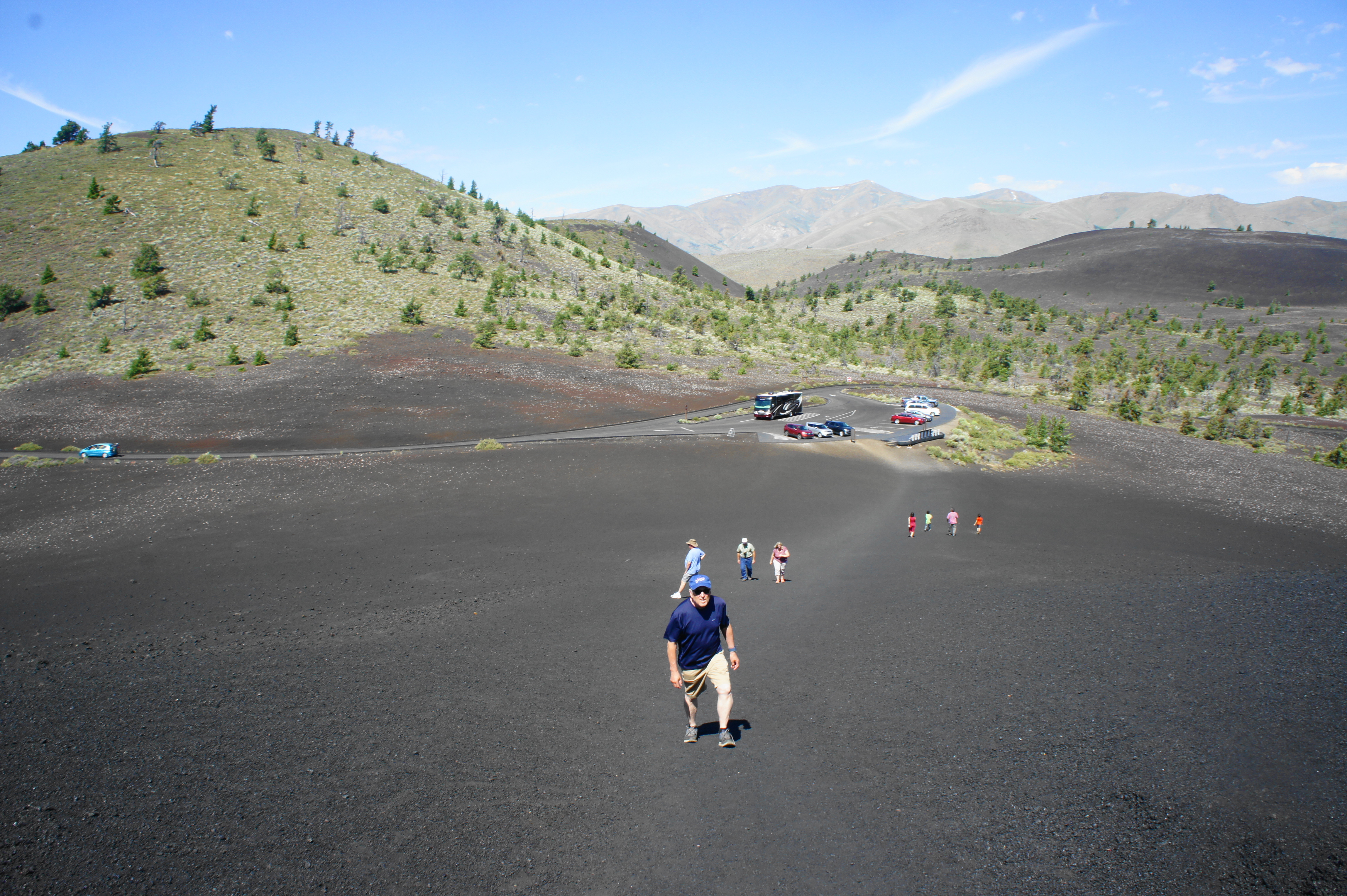 A real photograph of hikers climbing the steep cinder slope of Inferno Cone at Craters of the Moon National Monument in Idaho, with black lava fields stretching across the background under a bright blue sky