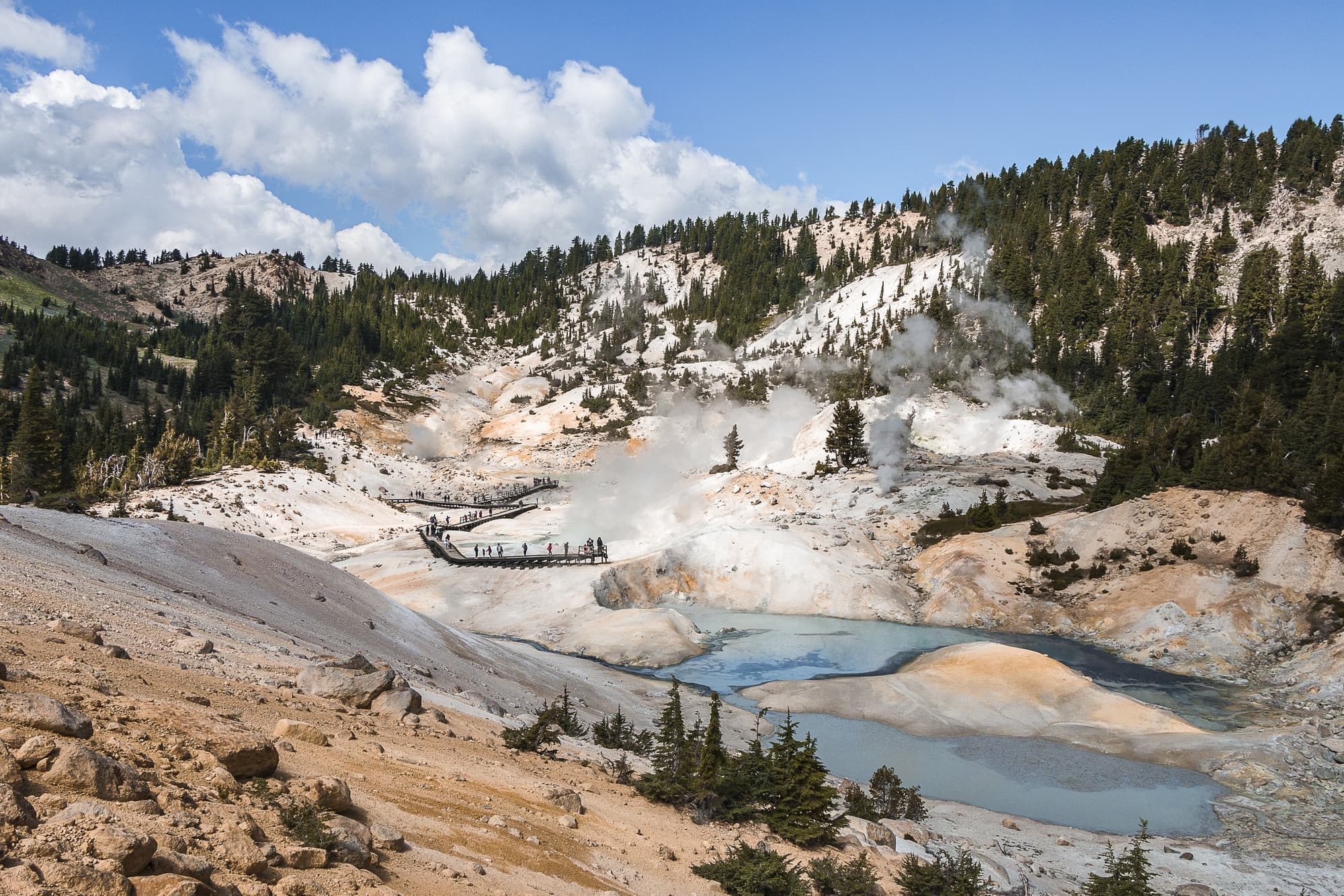 A real photograph of hikers getting ready at a Lassen Volcanic National Park trailhead on a sunny summer day, with backpacks, pine trees, and distant volcanic slopes in the background