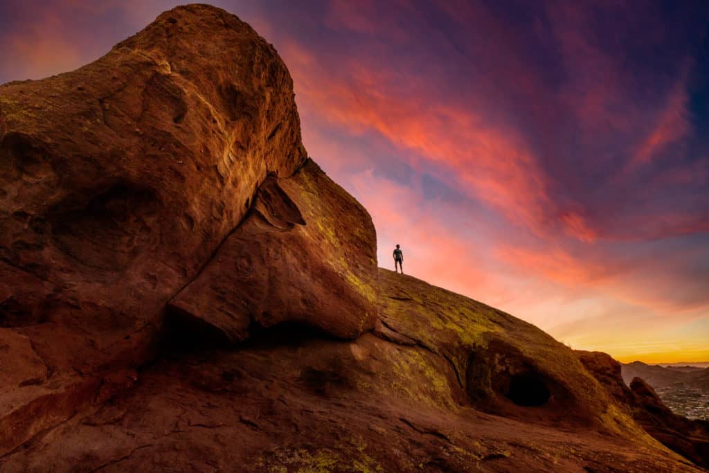 A real photograph of hikers on a rocky trail at sunrise on Camelback Mountain in Phoenix, with the city skyline hazy in the distance
