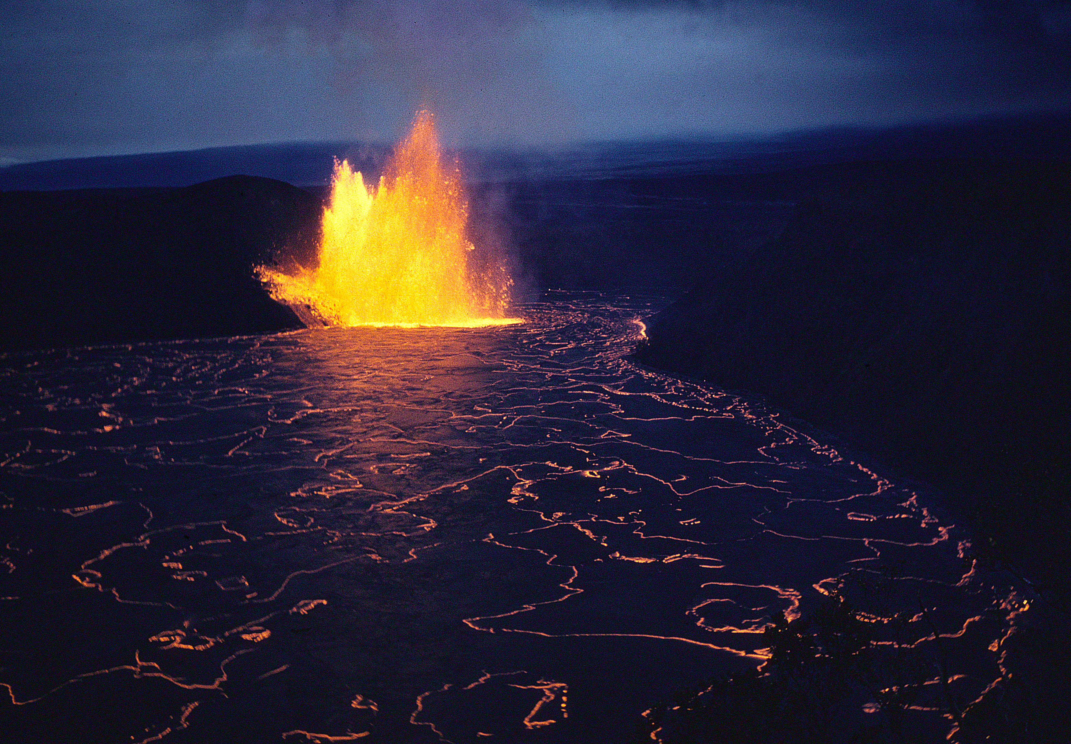 A real photograph of hikers walking across the cracked black surface of the Kīlauea Iki lava lake, surrounded by green rainforest walls and misty air