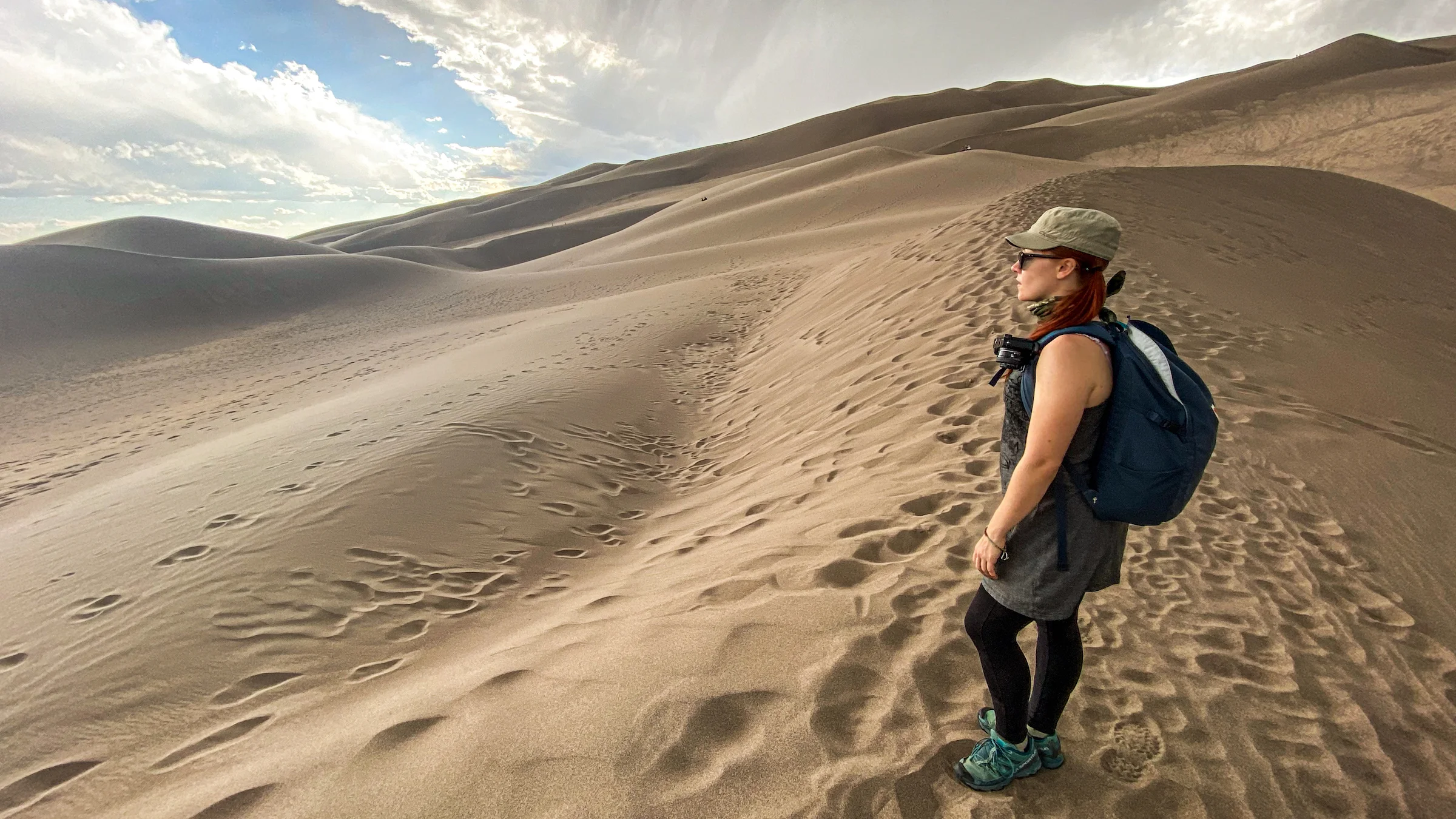 A real photograph of hikers walking along a high sand ridge at Great Sand Dunes National Park with the Sangre de Cristo Mountains in the background