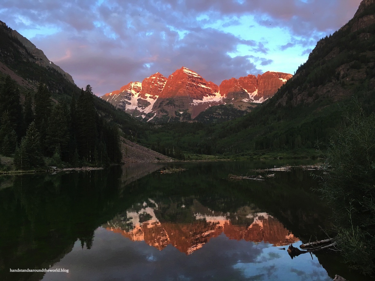 A real photograph of hikers walking along the Maroon Lake shoreline trail in the morning with the Maroon Bells peaks rising in the background