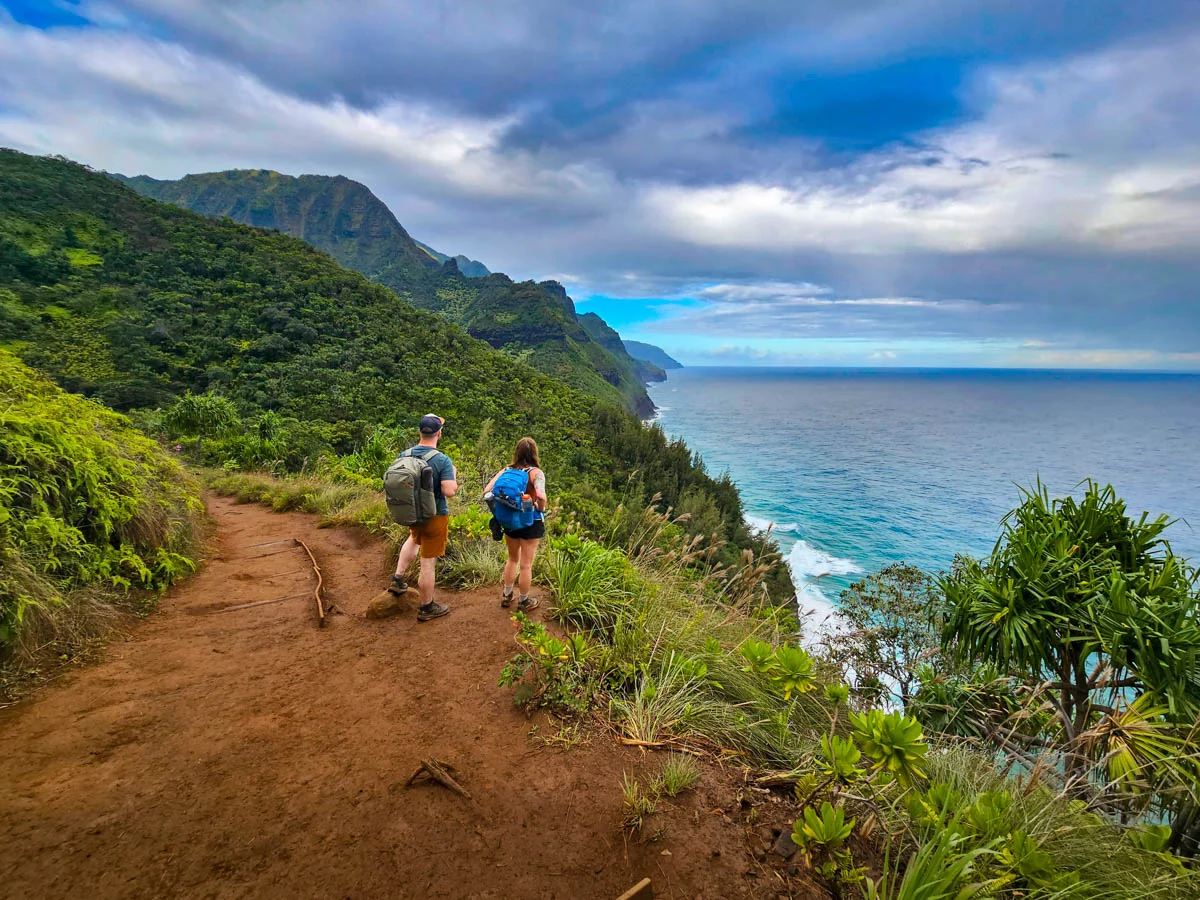 A real photograph of hikers walking along the narrow cliffside section of the Kalalau Trail with the ocean below and lush green slopes above