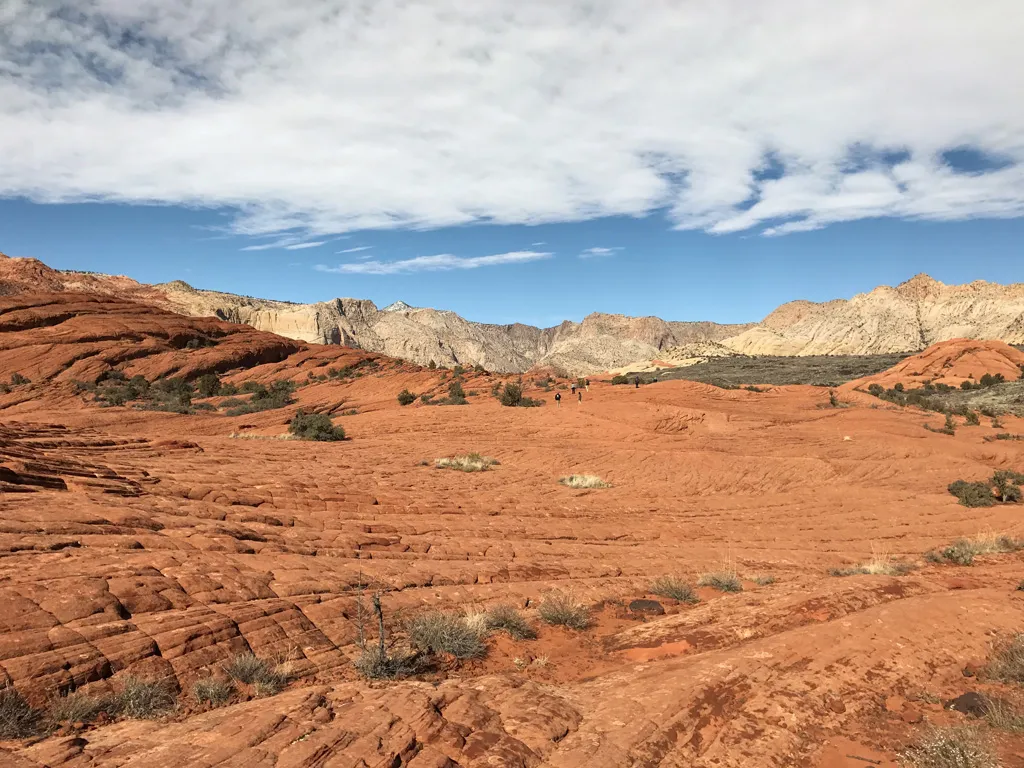 A real photograph of hikers walking along the petrified sand dunes in Snow Canyon State Park in Utah, with rippled sandstone and red cliffs under a clear blue sky