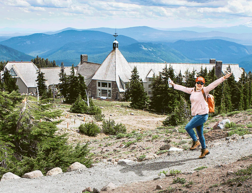 A real photograph of hikers walking on a dusty alpine trail above Timberline Lodge with Mount Hood rising in the background under a clear summer sky