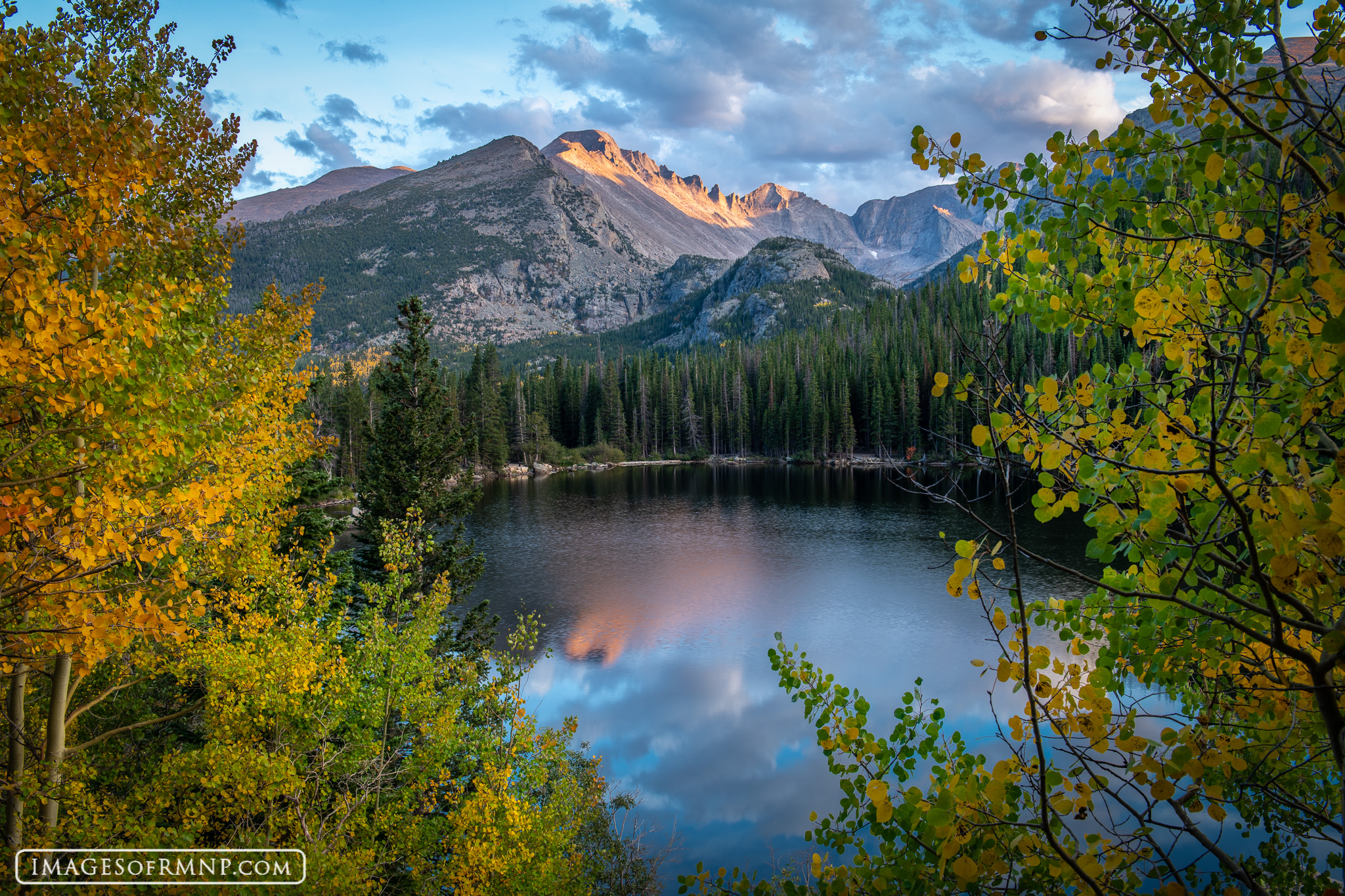 A real photograph of hikers walking on the Bear Lake loop trail through evergreen forest with rocky peaks rising in the background