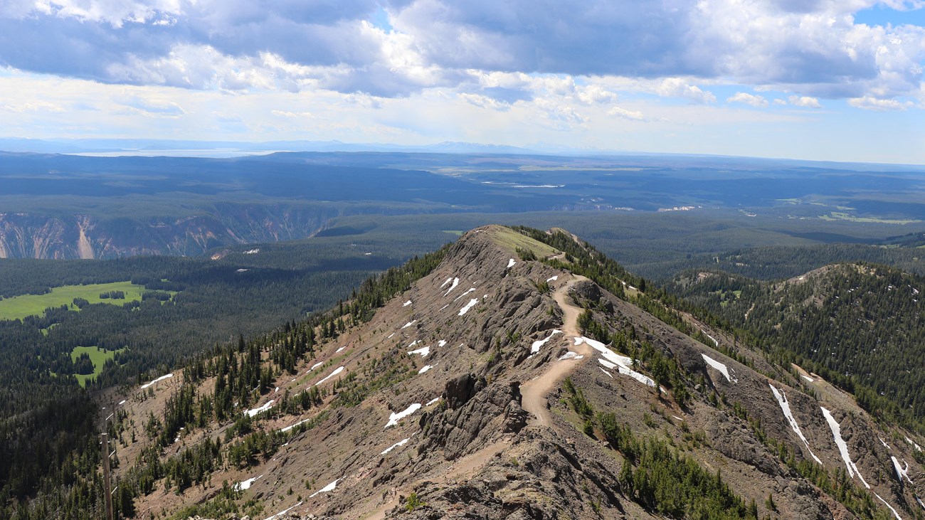 A real photograph of hikers walking on the Mount Washburn trail in Yellowstone on a clear summer day, with a wide alpine ridgeline, distant peaks, and open sky