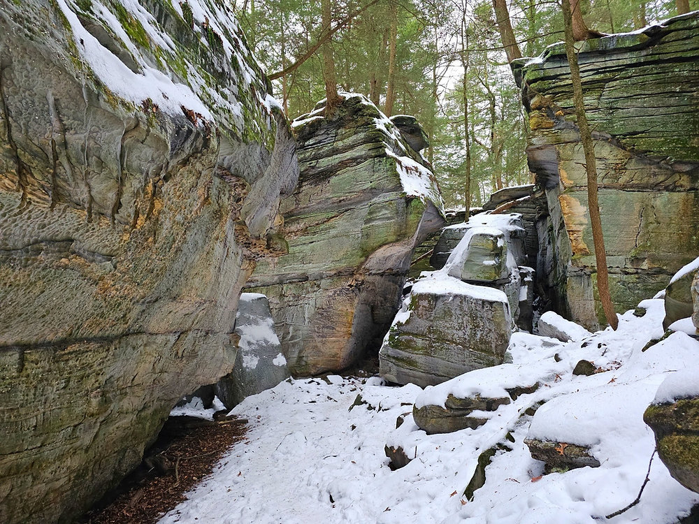 A real photograph of hikers walking through a narrow sandstone ledge corridor on the Ledges Trail, with ferns and moss along the rock walls