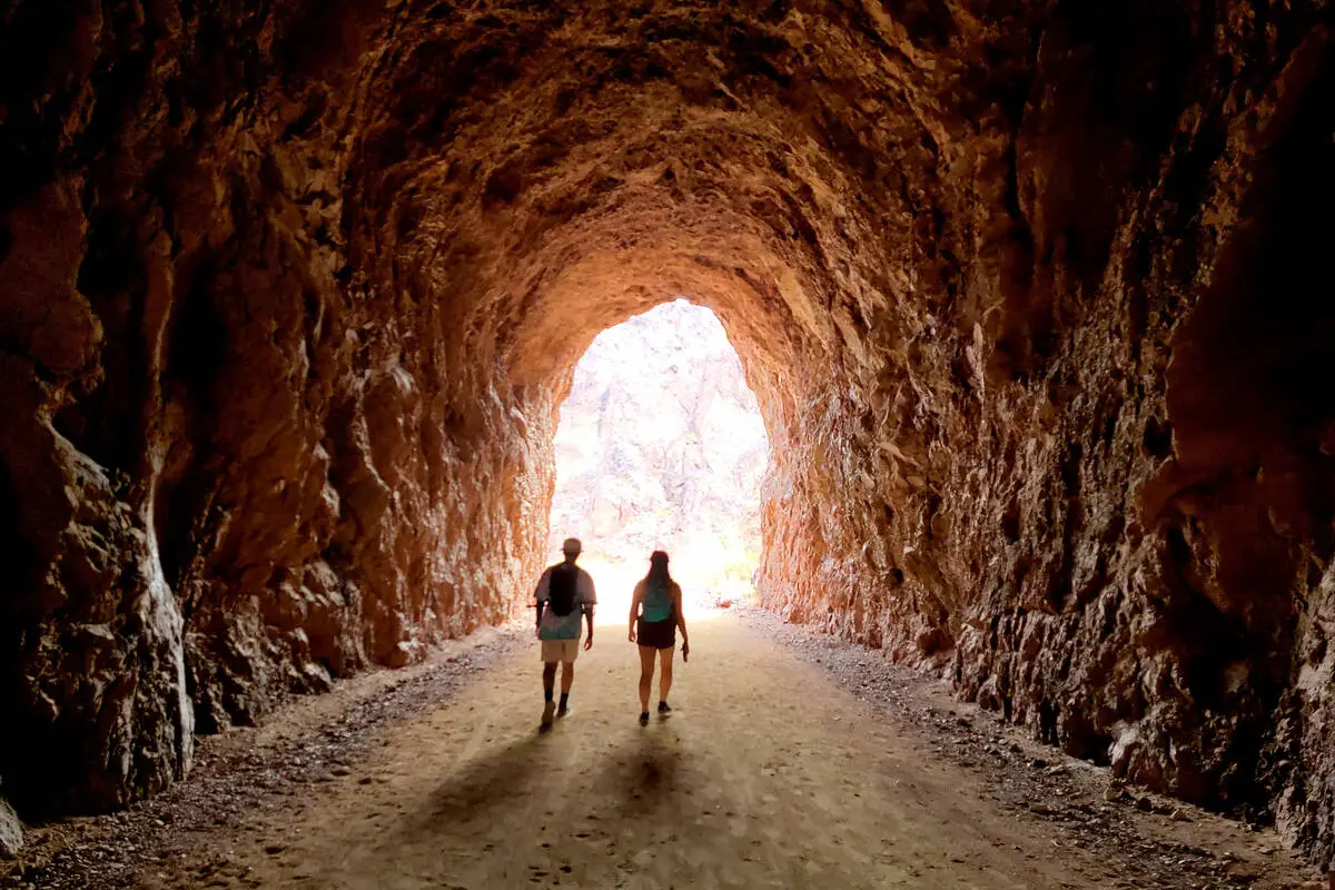 A real photograph of hikers walking through a wide, dark stone tunnel on the Historic Railroad Trail with bright desert light framing Lake Mead in the distance