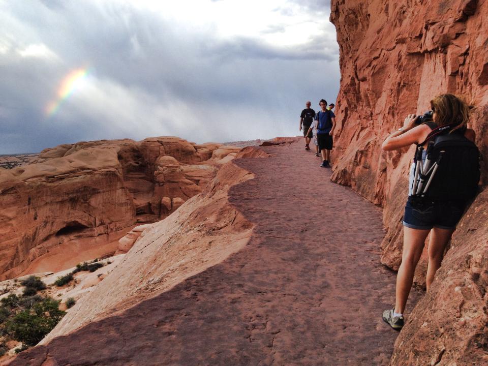 A real photograph of hikers walking up the Delicate Arch trail on wide slickrock slopes with red sandstone and desert shrubs under a clear blue sky