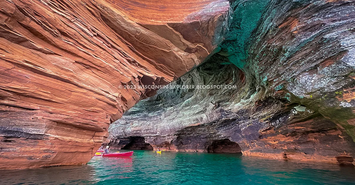A real photograph of kayakers floating near the sandstone sea caves at Meyers Beach in the Apostle Islands, with clear summer light reflecting off Lake Superior and cave openings visible in the cliff
