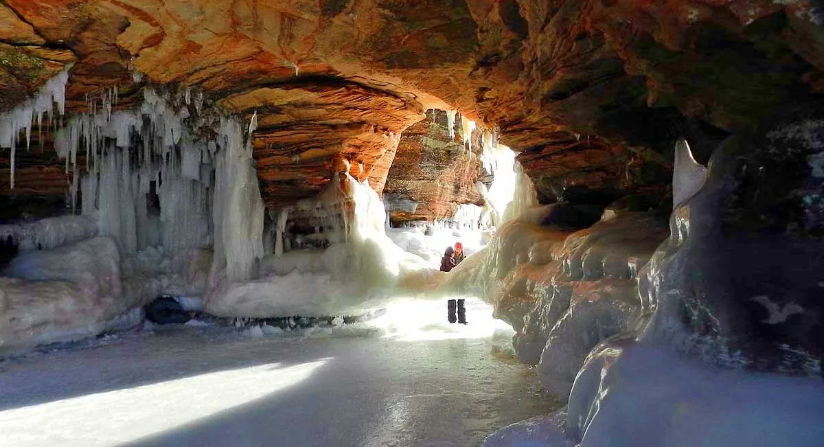 A real photograph of people walking near tall blue-white ice formations along the Lake Superior shoreline at Meyers Beach, with frozen spray ice clinging to sandstone cliffs on a bright winter day