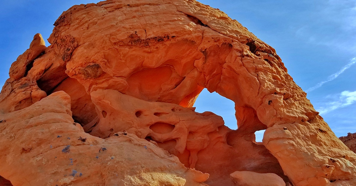 A real photograph of red sandstone formations in Valley of Fire State Park, Nevada, with a lone hiker on a sandy trail under bright midday sun