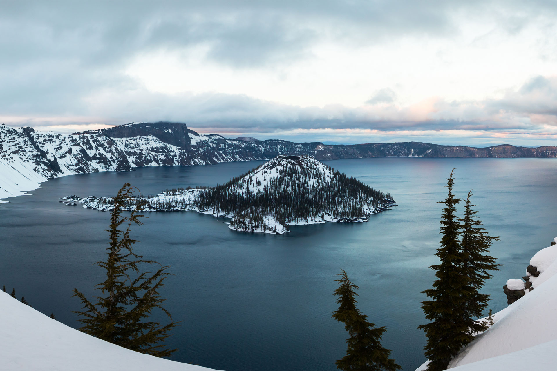 A real photograph of snowbanks lining Rim Drive with a viewpoint turnout and Crater Lake visible beyond, showing winter or early spring conditions