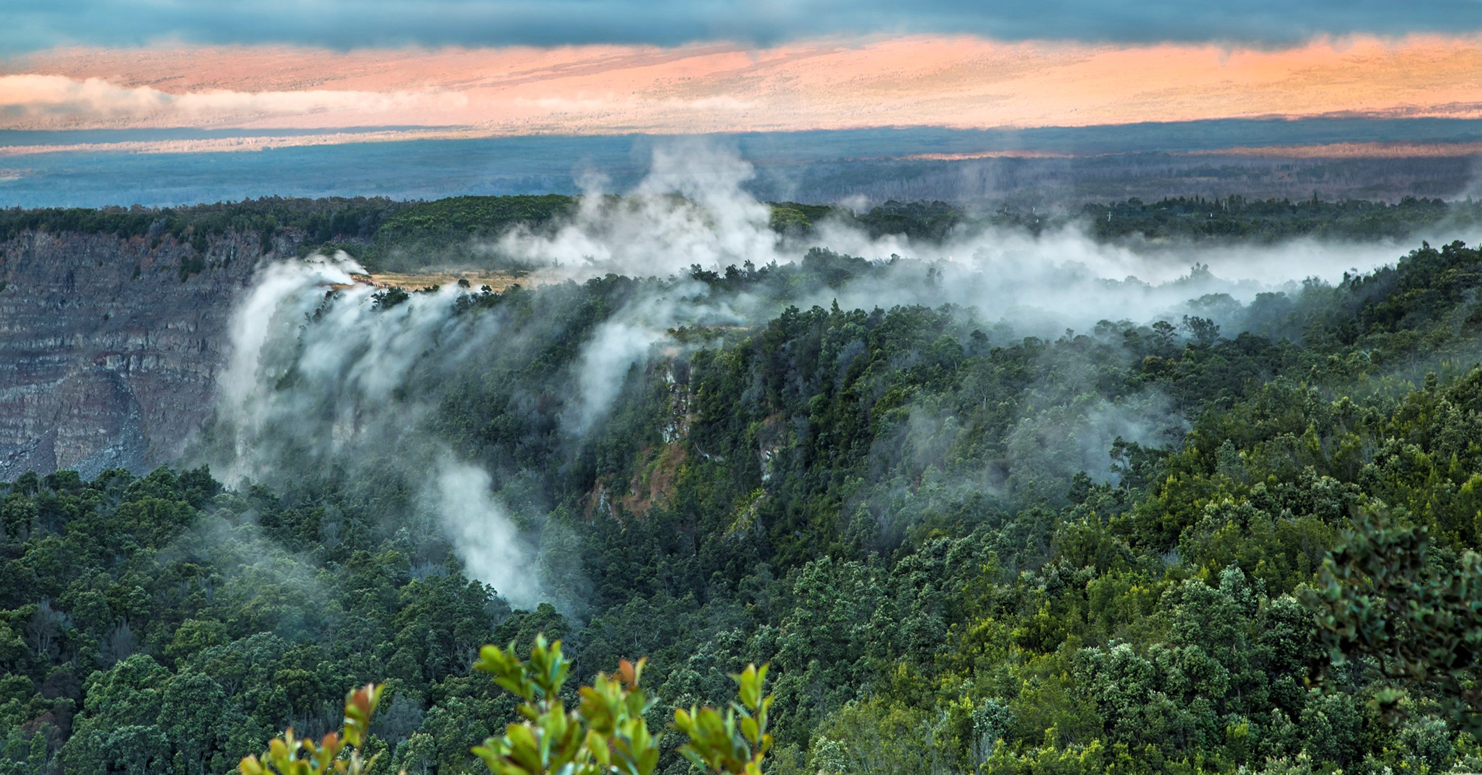 A real photograph of steam rising from the Sulphur Banks area in Hawaiʻi Volcanoes National Park, with a boardwalk path cutting through pale mineral-stained ground and low fog