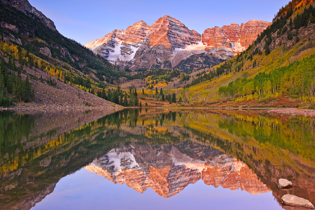 A real photograph of sunrise light hitting the Maroon Bells peaks reflected in the still water of Maroon Lake with a few visitors standing on the shoreline