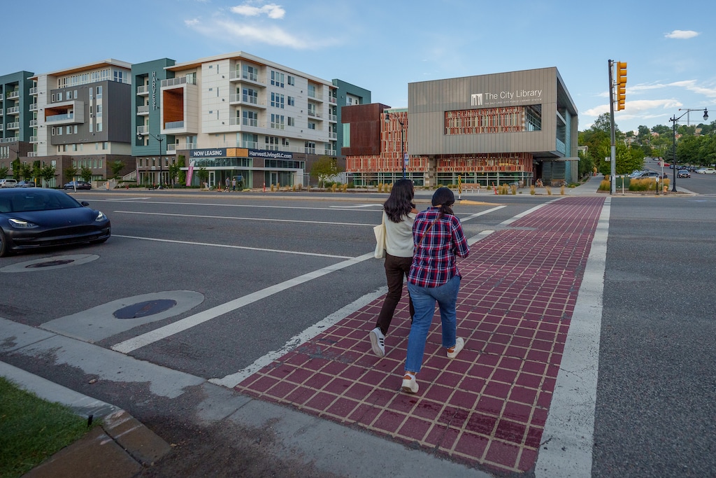 A real photograph of the 9th and 9th neighborhood in Salt Lake City with small storefronts, pedestrians crossing, and leafy trees along the sidewalk