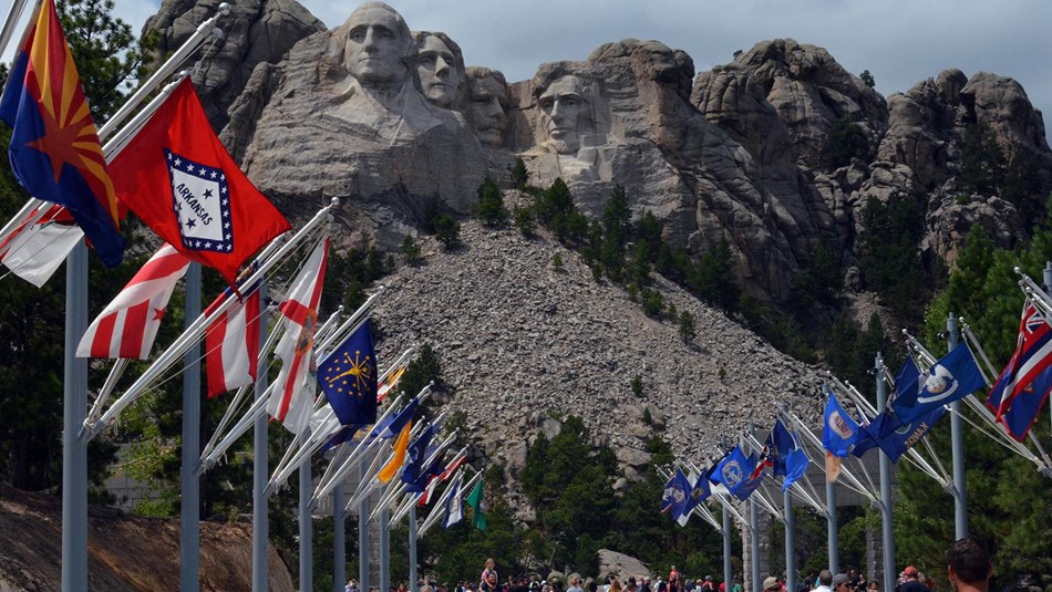 A real photograph of the Avenue of Flags at Mount Rushmore in the morning with flags lining the walkway and the memorial visible in the distance