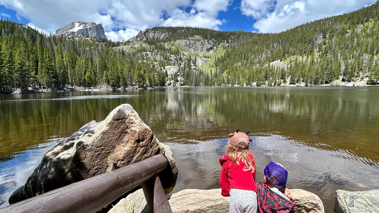 A real photograph of the Bear Lake Trailhead area in Rocky Mountain National Park on a bright summer morning, with pine forest, distant peaks, and hikers preparing near the trailhead signs