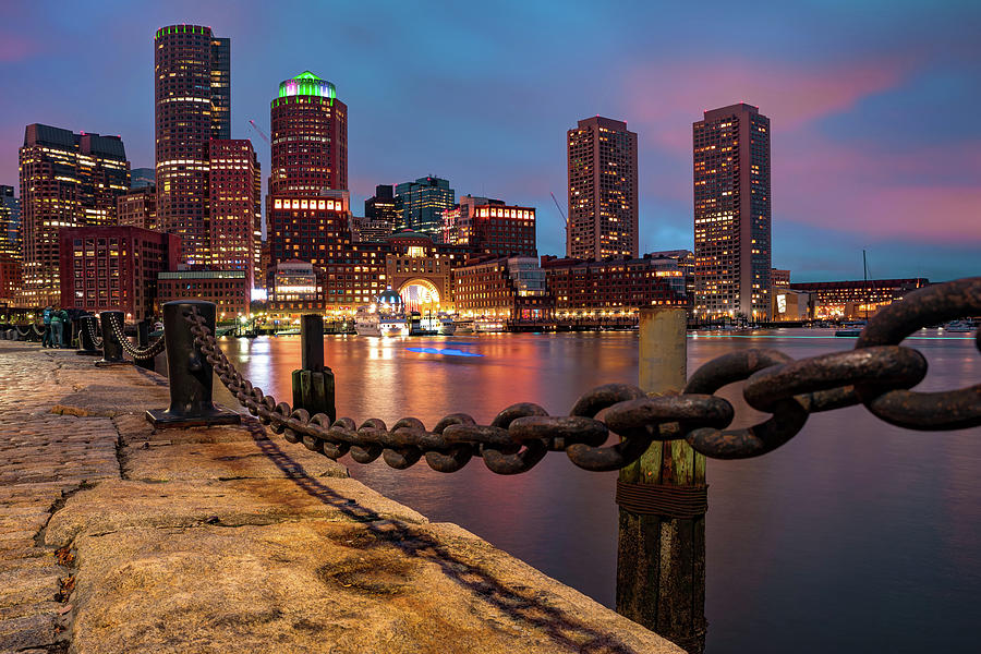 A real photograph of the Boston skyline viewed across the harbor at sunset with calm water and boats in the foreground