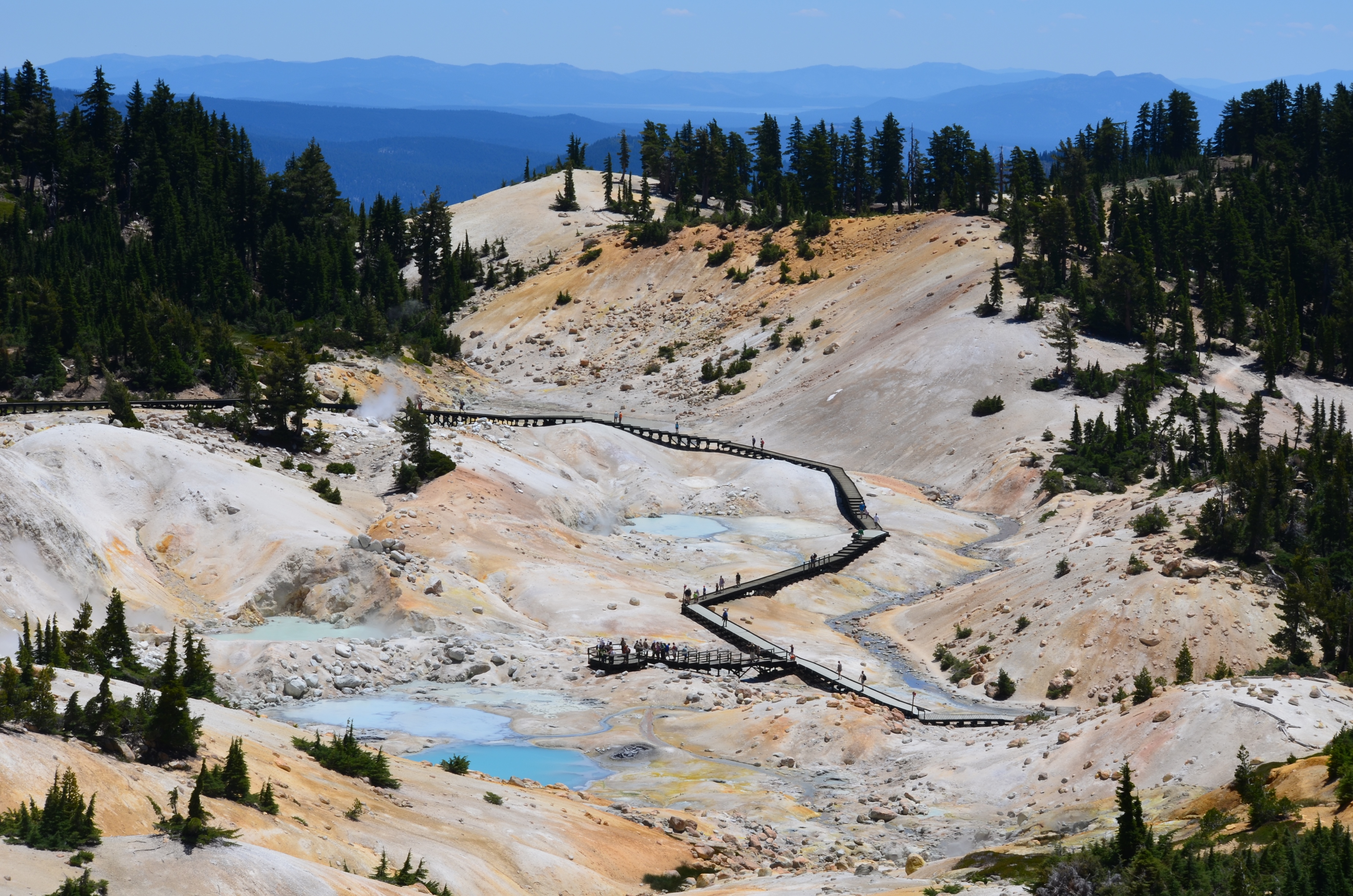 A real photograph of the Bumpass Hell boardwalk in Lassen Volcanic National Park with steam rising from hydrothermal vents and hikers walking along the wooden path on a clear summer day