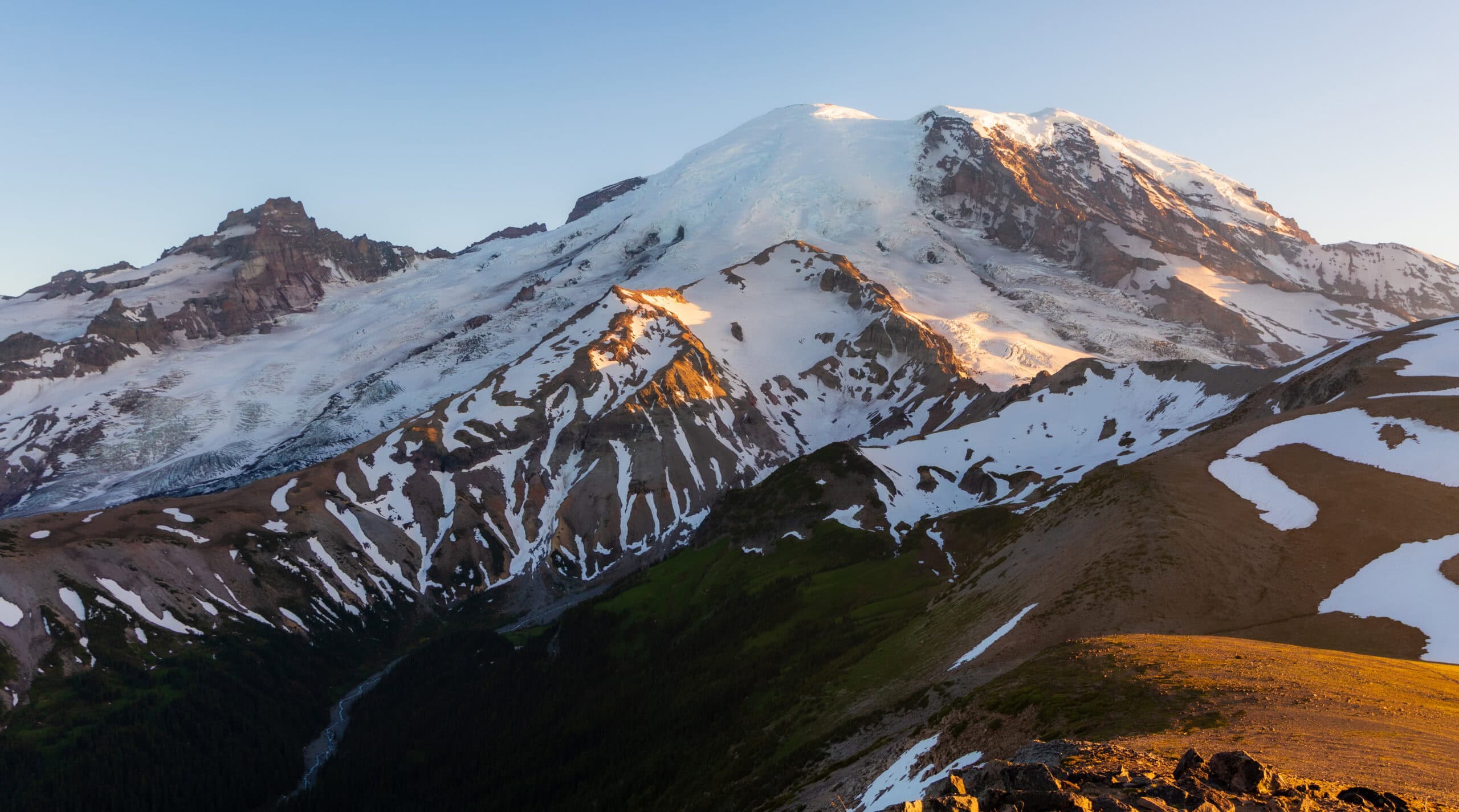 A real photograph of the Burroughs Mountain trail near Sunrise with rocky tundra, distant hikers on the ridgeline, and Mount Rainier dominating the background
