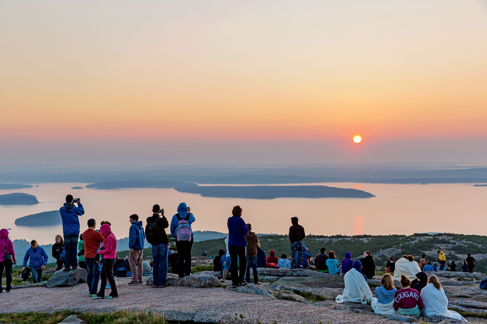 A real photograph of the Cadillac Mountain summit at sunrise in Acadia National Park, with pink light over the Atlantic Ocean and a small crowd of visitors standing near the granite overlook