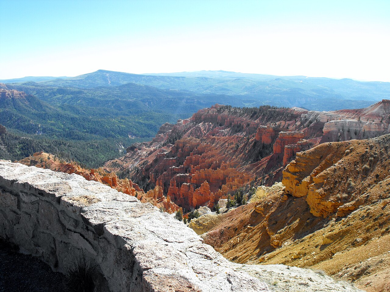 A real photograph of the Cedar Breaks amphitheater viewed from the rim on a clear summer day, showing red-orange hoodoos and cliffs dropping into a wide basin with evergreen forest in the distance