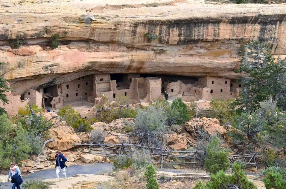 A real photograph of the Chapin Mesa Archaeological Museum area at Mesa Verde, with stone walls, desert plants, and visitors walking between exhibits on a sunny day