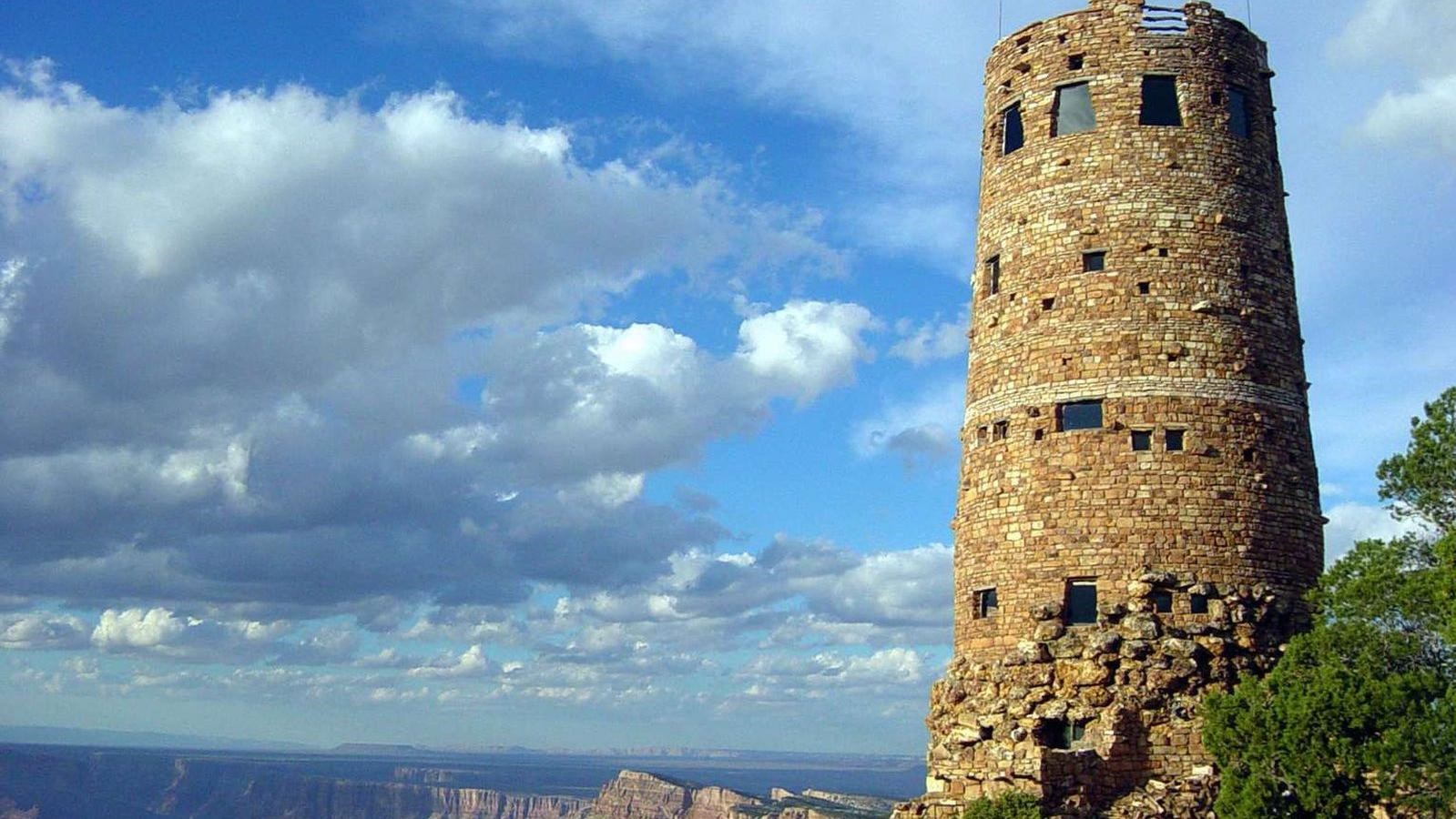 A real photograph of the Desert View Watchtower on the Grand Canyon South Rim, with the stone tower rising above the rim and the canyon stretching behind it under a clear blue sky