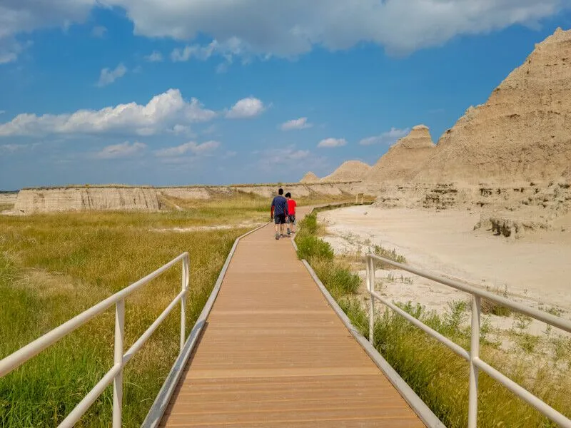 A real photograph of the Door Trail boardwalk in Badlands National Park in the early morning, leading toward jagged rock formations under a pale pink sky